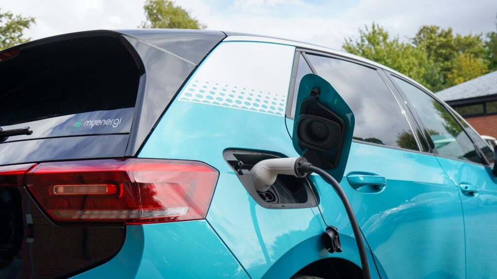 A turquoise electric car, perfect for those buying a new car, is parked and plugged into a charging point. Its charging port flap is open with the cable connected. Trees and a building can be seen in the background.