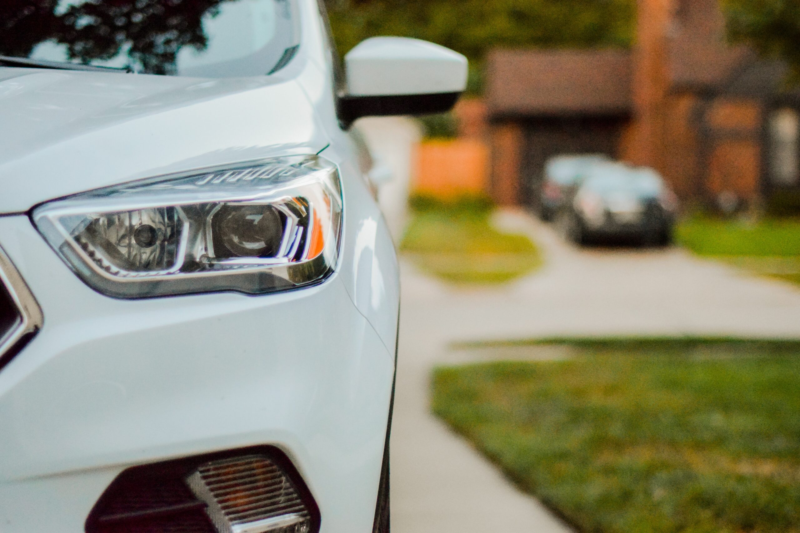Close-up of the front left side of a white car parked on a drive—perfect for anyone considering buying a new car—with a suburban house, green lawn, and another car blurred in the background.