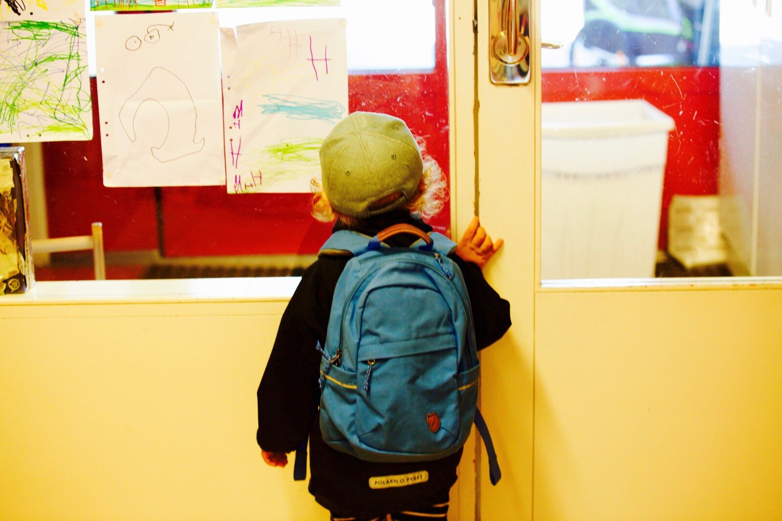 A young child with a blue rucksack and a green cap stands facing a closed door, looking through the glass. Colourful children's drawings are displayed on the door.