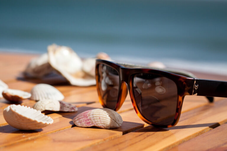 A pair of sunglasses rests on a wooden surface surrounded by seashells, with the blurred background suggesting a beach or seaside setting.