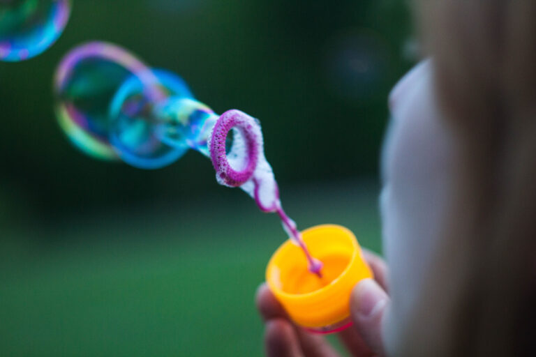 A close-up of a person blowing soap bubbles outdoors, with a bubble wand dipped in a yellow container. Several colourful bubbles are visible against a blurred green background.
