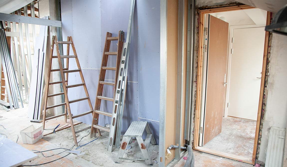 A room undergoing refurbishment with ladders, building materials, and tools scattered about. An open door reveals another unfinished space beyond, prompting thoughts of where to buy building supplies for projects like this. Exposed pipes and plasterboard panels are visible.