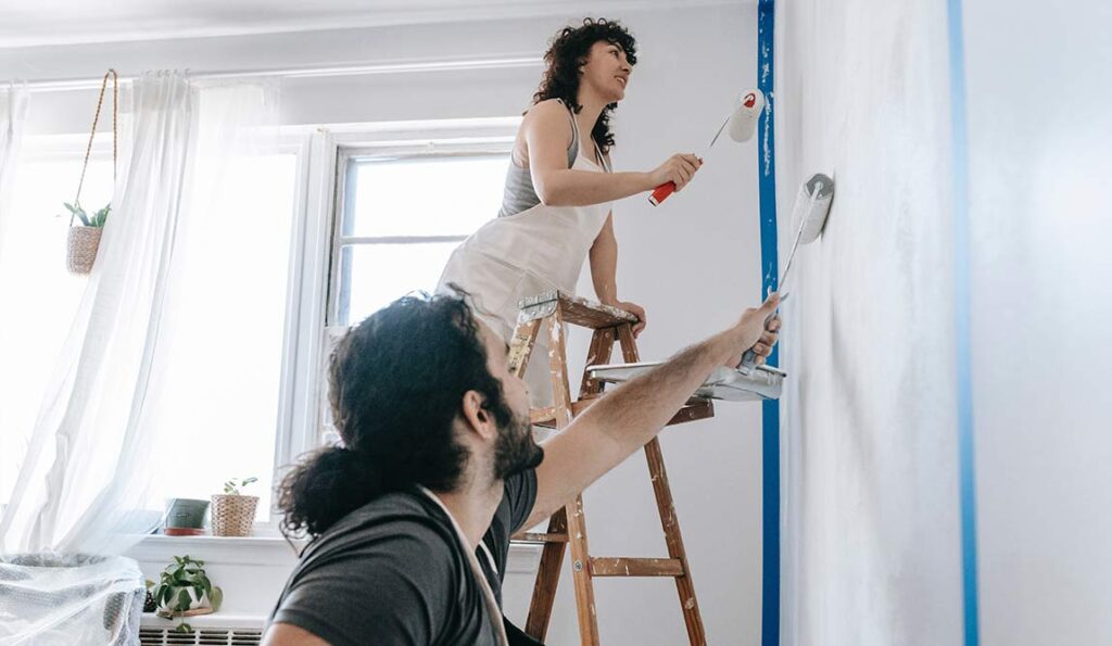 A woman stands on a wooden ladder painting a wall with a roller, whilst a man below also paints. Sunlight streams through the window onto plants on the sill—perhaps they’re wondering where to buy building materials for their project.