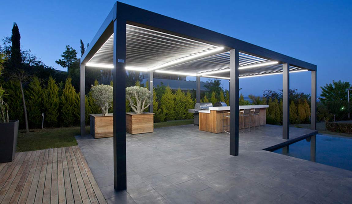 Modern outdoor patio with a sleek pergola featuring built-in lighting, a bar area with stools, large potted plants, and surrounding greenery. Pergolas with retractable roofs add versatility to the space, which is captured at dusk under a clear blue sky.