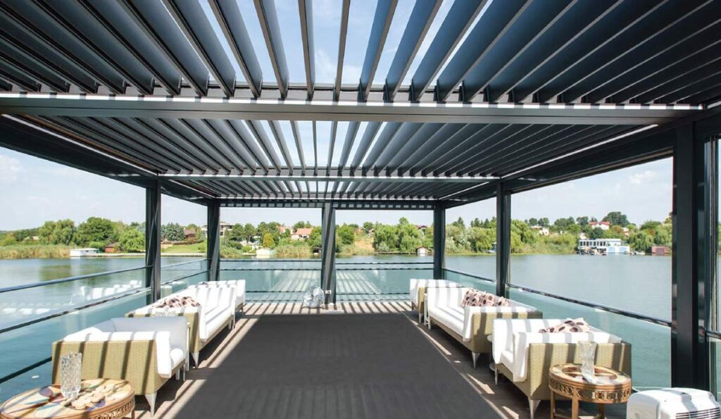 Modern outdoor lounge with white sofas under pergolas with retractable roofs, overlooking a calm lake and green trees on a sunny day.