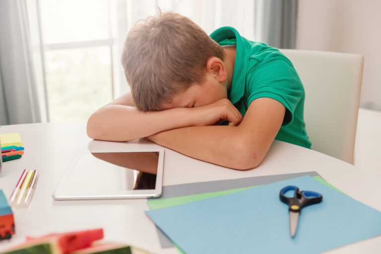 A young boy in a green shirt sits at a table with his head resting on his arms, looking tired or frustrated. On the table are an electronic tablet, coloured paper, scissors, pens, and a pencil.
