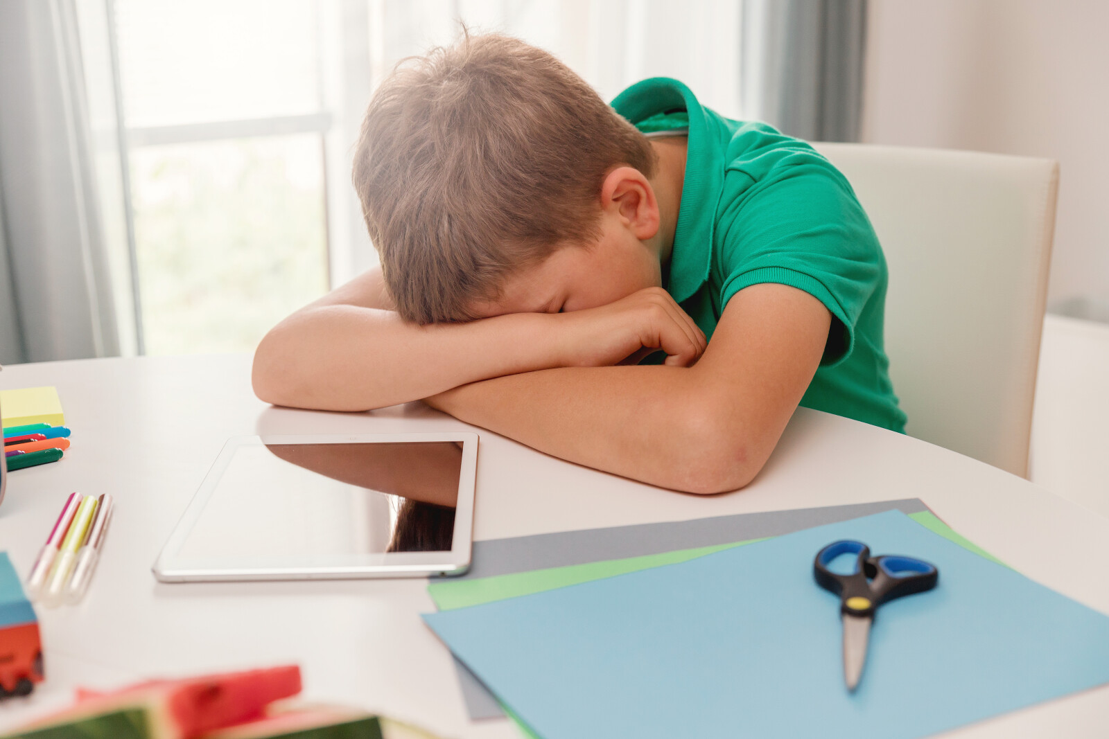 A young boy in a green shirt sits at a table with his head resting on his arms, looking tired or frustrated. On the table are an electronic tablet, coloured paper, scissors, pens, and a pencil.