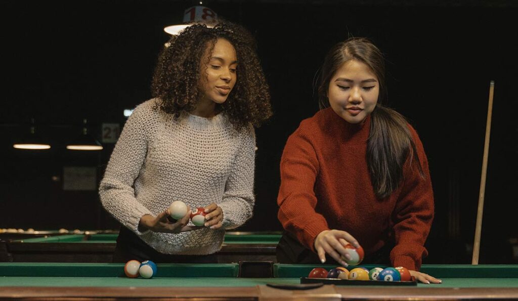 Two women enjoy their hobby next to a pool table, arranging billiard balls. One wears a light jumper and holds balls, while the other, in red, places them on the table. The dimly lit background features hanging lights.