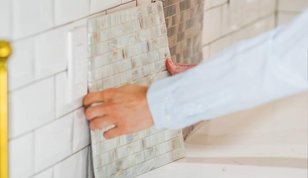 A person installing a sheet of mosaic tile splashback on a kitchen wall near a white electrical socket. Their hands position the tile, part of a light blue sleeve visible—wondering where to buy building materials for your own project?.