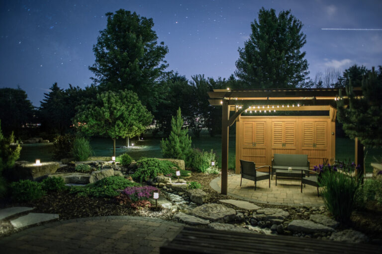 A back garden patio at night features a wooden pergola with chairs and a table, surrounded by landscaped gardens and stone paths, softly lit by garden lights under a starry sky.