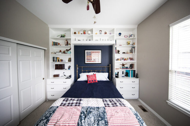 A neatly organised bedroom with a bed centred against built-in white shelves filled with books, toys, and decor. An American flag artwork hangs above the bed. Sunlight streams in from a window on the right.