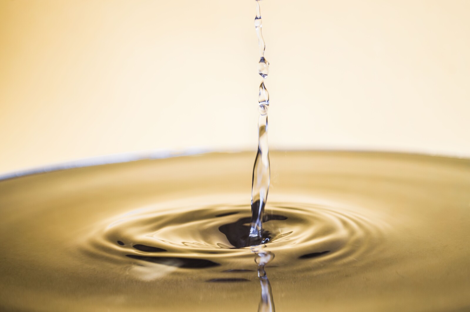 Close-up of a stream of water falling into a calm, shallow pool, creating gentle ripples on the surface, with a soft yellow background.