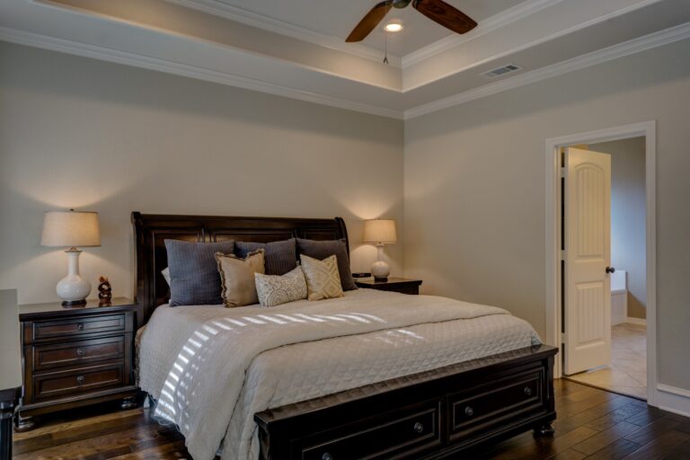 A cosy bedroom with a large dark wood bed, beige bedding, decorative cushions, matching bedside tables with lamps, a ceiling fan, and an open door leading to a bright adjoining room.