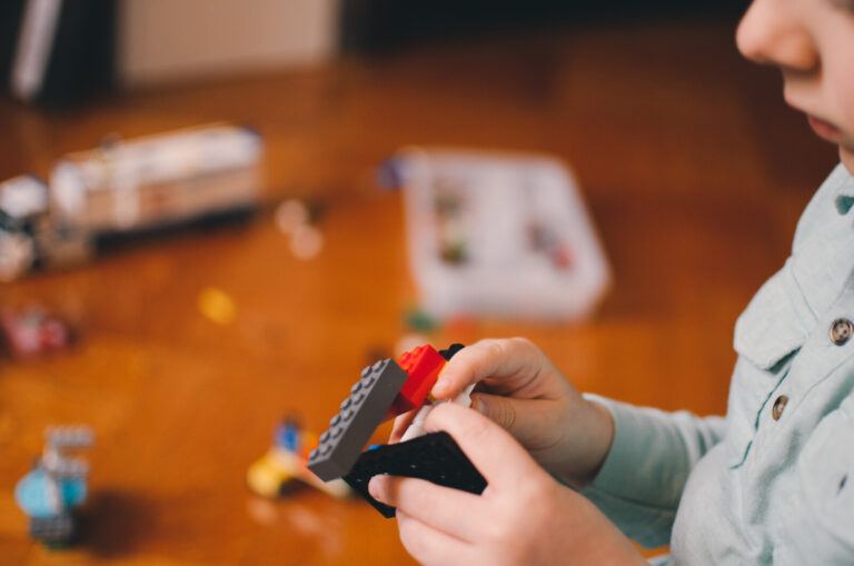 A child in a light blue shirt is building with colourful LEGO bricks, holding pieces in both hands. The background is out of focus, showing more LEGO bricks and toys on a wooden surface.