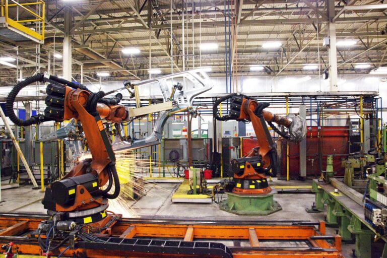 Two industrial robotic arms work on assembling a car body, producing sparks in a brightly lit, modern factory setting with metal structures and machinery in the background.