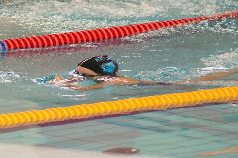 A swimmer wearing a black swimming cap and goggles swims in a lane of an indoor pool, bordered by yellow and red lane markers.
