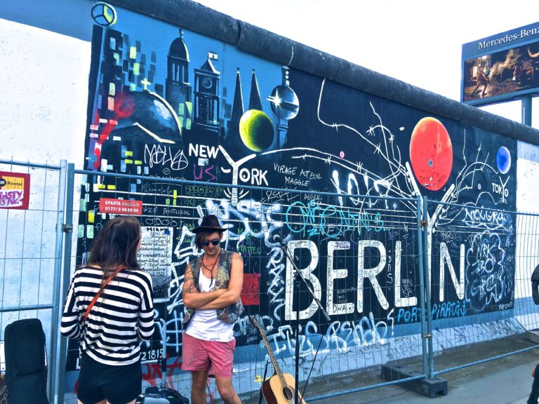 Two people stand in front of the Berlin Wall, covered in colourful graffiti and “BERLIN.” A guitar rests by the mural and a metal fence is nearby—perfect for those exploring The Best German Restaurants in Berlin after taking in cityscape art.