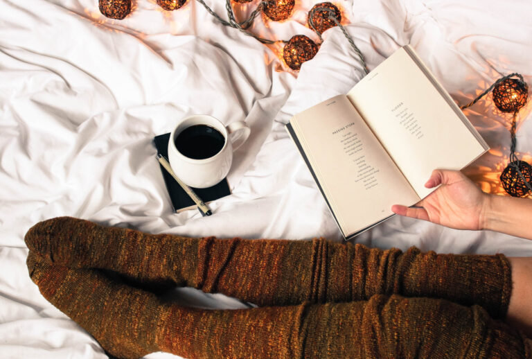 A person wearing cosy brown socks sits on a white bed with fairy lights, holding an open book. Nearby are a cup of coffee, a notebook, and a pen, creating a warm and relaxing atmosphere.
