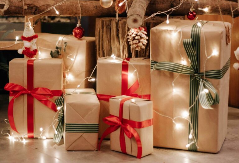 A group of wrapped Christmas presents with red and green ribbons sits under a tree, surrounded by festive fairy lights and baubles—perfect gift ideas for adults 2022.