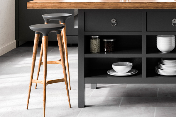 Two wooden barstools with grey seats sit by a dark kitchen island with open shelves displaying dishes and jars—a stylish example of 2023 kitchen touch-ups to consider, set on a light tiled floor.