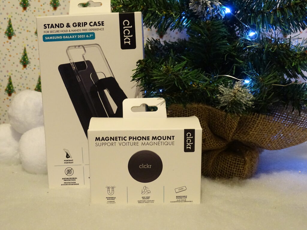 Two Clckr products—a Stand & Grip Case for Samsung Galaxy and a Magnetic Phone Mount—sit on a white surface in front of a decorated Christmas tree, making perfect gift ideas for adults 2022. Blue lights and artificial snow add festive charm.