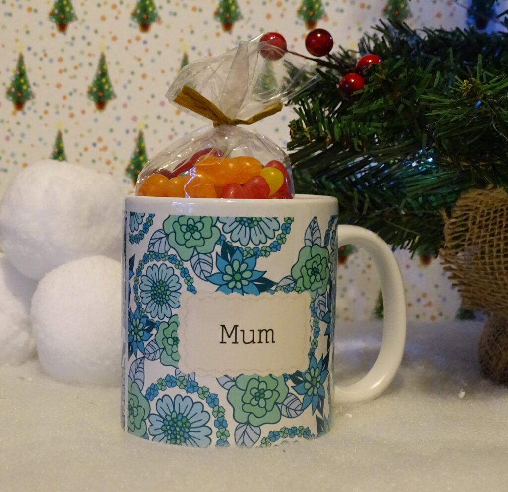 A white mug with blue floral patterns and the word "Mum" printed on it holds colourful jelly beans. Set on fake snow near a small artificial Christmas tree and snowballs, it's one of the sweetest gift ideas for adults 2022.