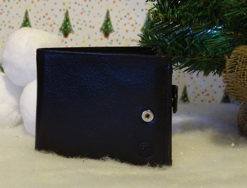 A black leather wallet sits on artificial snow next to a small Christmas tree and white snowballs, making it one of the perfect gift ideas for adults 2022 against a festive backdrop with tiny green Christmas trees.