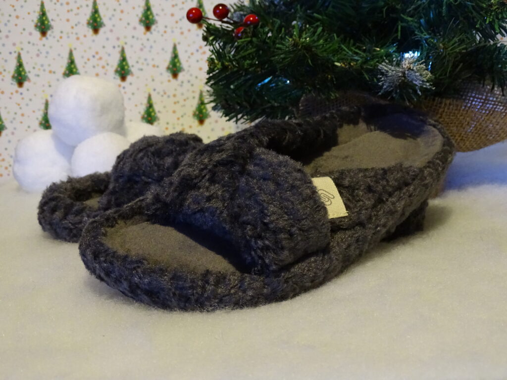 A pair of fluffy, dark grey slippers rests on a white surface beside a decorated Christmas tree—perfect gift ideas for adults 2022—with snowball decorations in the background.