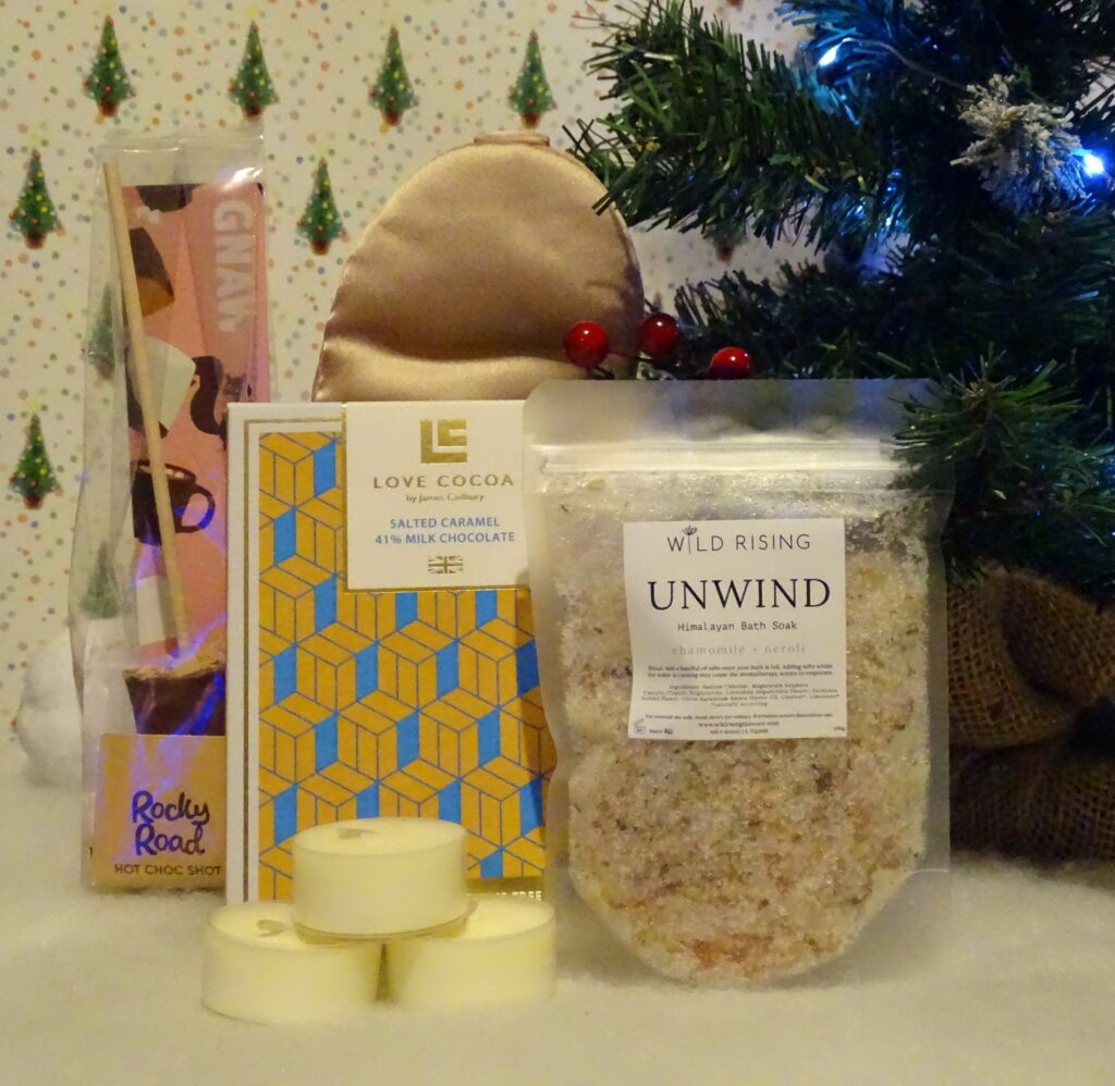 A festive arrangement features a Rocky Road hot chocolate stirrer, a Love Cocoa salted caramel chocolate bar, a Wild Rising Unwind bath soak, and three tealight candles—perfect gift ideas for adults 2022—set before a Christmas tree and stocking.
