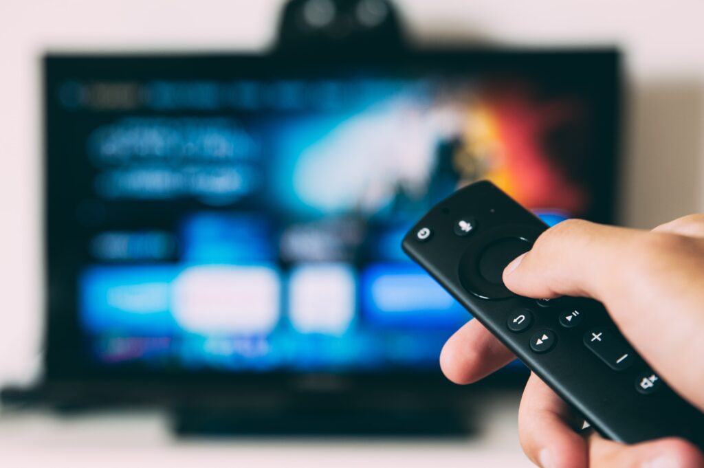 A hand holding a remote control points towards a blurred television screen displaying a streaming service interface with colourful icons and options, perfect for browsing educational content and tips on how to boost your child's education.