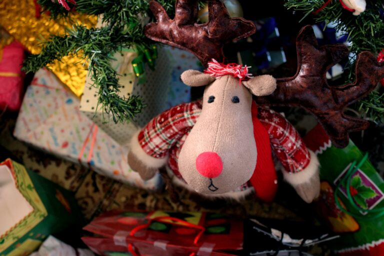 A plush reindeer toy with a red nose and checked jumper sits under a decorated Christmas tree, surrounded by colourful wrapped Christmas presents for children 2022.