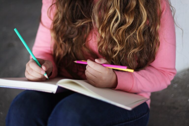 A person with long, wavy hair wearing a pink jumper is writing in an open notebook with a green pencil and holding a pink pencil, illustrating how to help your children with their homework in a calm and supportive way.