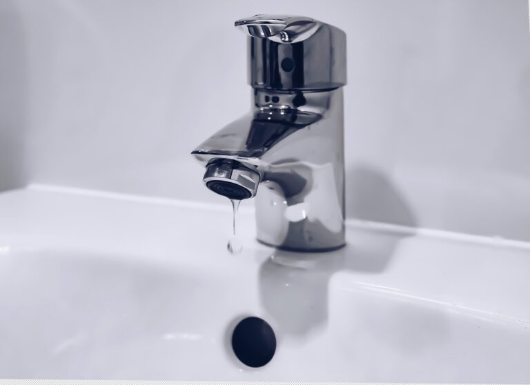 A close-up of a chrome tap over a white basin with a single drop of water dripping from the spout, highlighting the importance of bathroom efficiency. Discover tips to save money: Is your bathroom energy efficient? Cool, clean tones and blurred background.