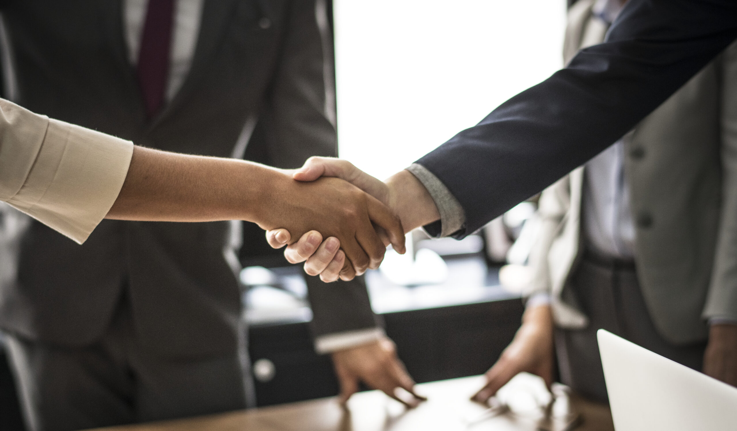 Two people in business attire are shaking hands across a table, suggesting a professional agreement or partnership—an example of how to keep your customers feeling helped and supported. Other individuals and a laptop are visible in the background.