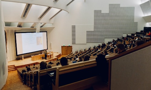 A modern lecture theatre with rows of wooden seats, several people seated, and a large projection screen at the front displaying a presentation on 3 Vital Steps To Plan For Your Child’s Higher Education. The room features white walls and a high ceiling with angled lights.