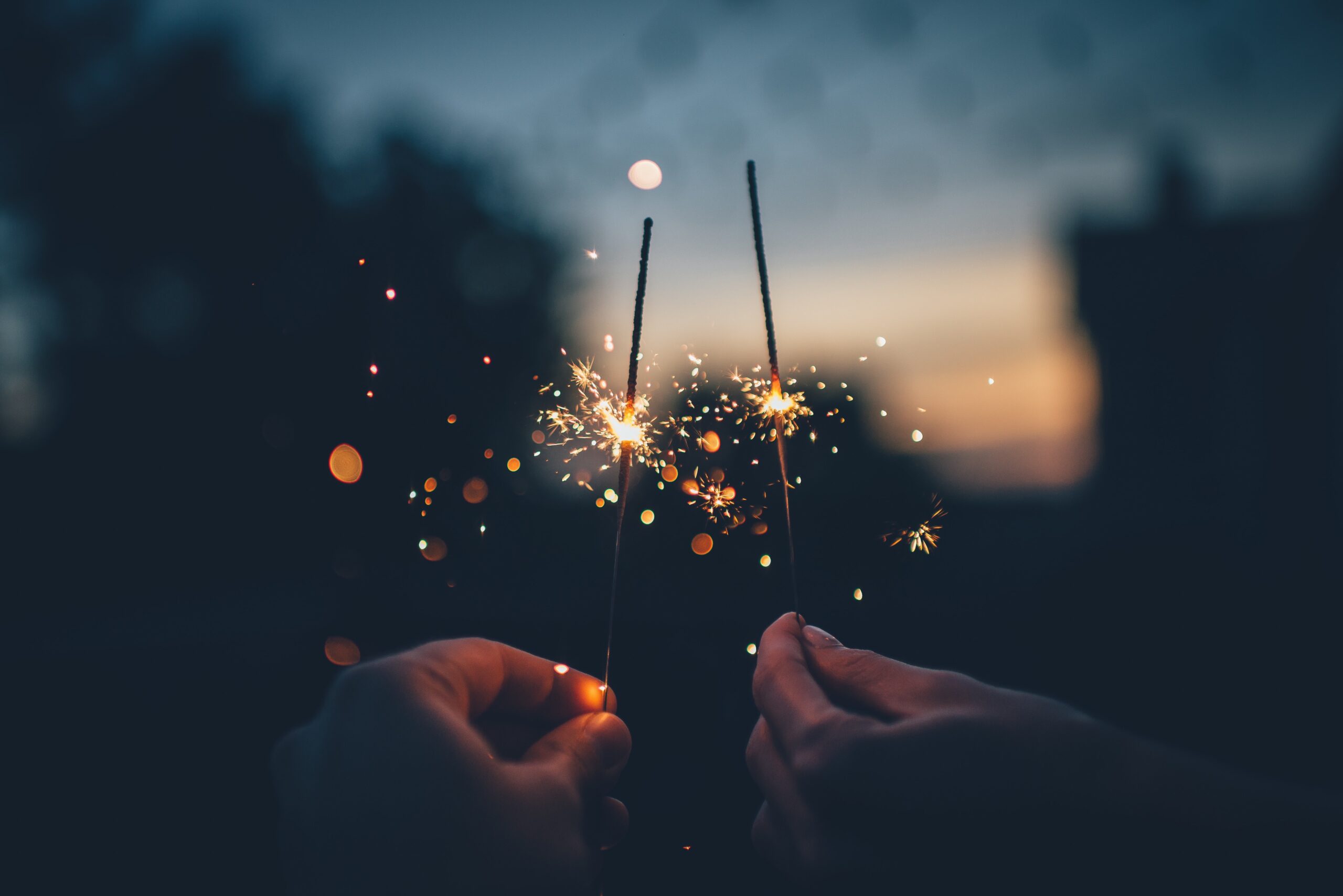 Two hands holding lit sparklers close together, with bright sparks and bokeh lights against a dark, blurred background at dusk—perfect for capturing magical moments or finding gift ideas to surprise your spouse on the wedding night.