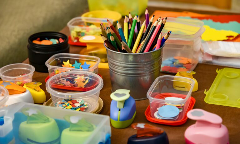 A table covered with art supplies highlights the importance of expressive art & design lessons in nursery schools, featuring coloured pencils in a metal cup, plastic containers with paper cut-outs, and various crafting punches in vibrant shapes and colours.