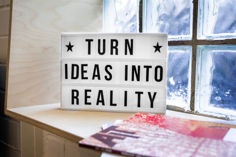 A lightbox sign on a wooden shelf reads "TURN IDEAS INTO REALITY," echoing the spirit of Startup Success: 5 Steps To Achieve Your Goals, placed next to magazines by a window with glass bricks.