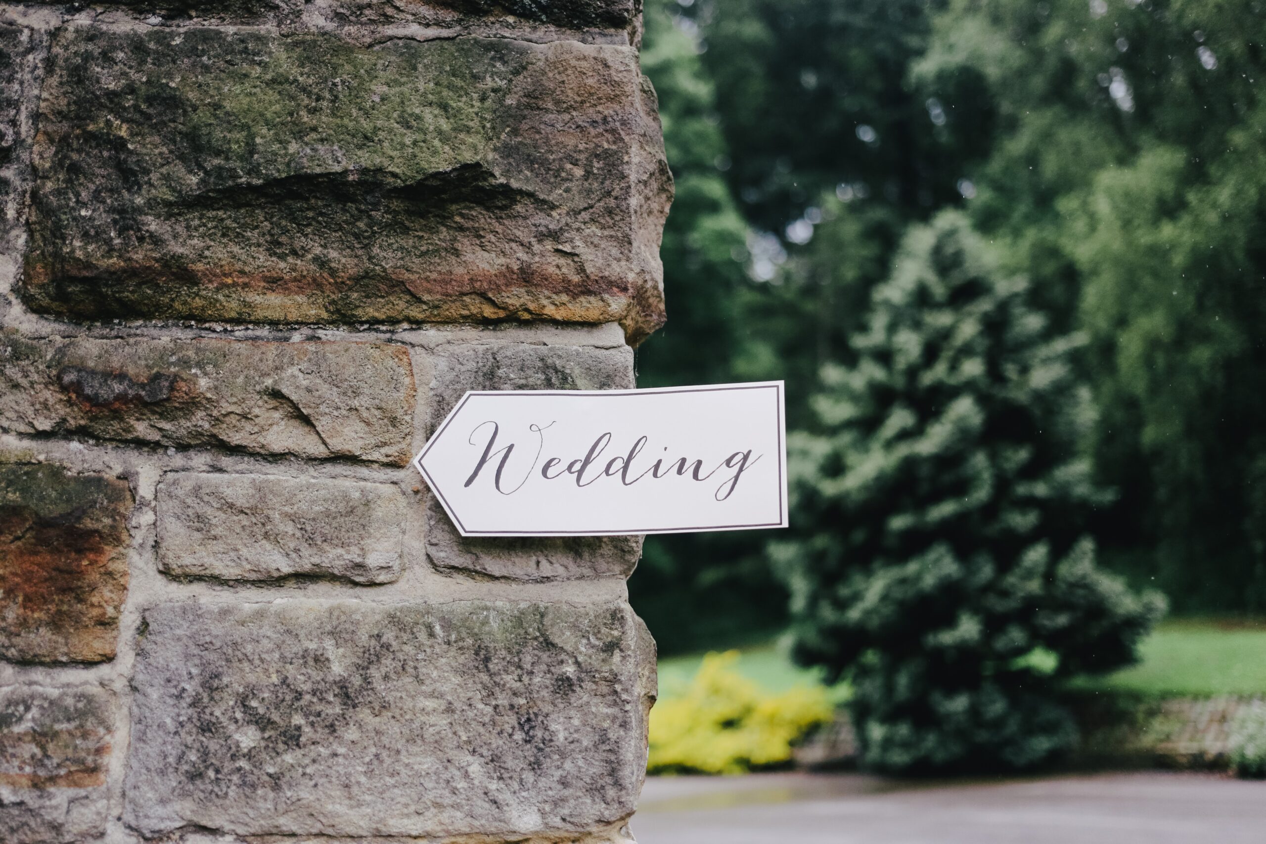 A white arrow-shaped sign with "Wedding" in cursive is attached to a stone wall, pointing right—reminding guests of the important details when planning your wedding. Green trees and bushes are visible in the blurred background.