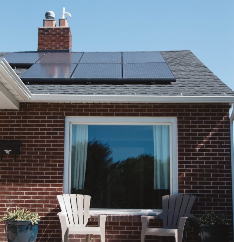 A brick house showcases the benefits of installing solar panels on your home, featuring rooftop panels, a large window with white curtains, and two empty patio chairs with a small table outside.