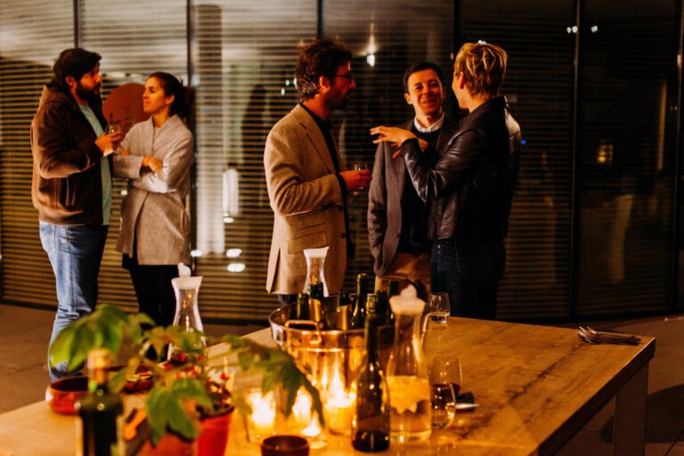 A group of people socialising at an indoor gathering, with two pairs engaged in conversation. A wooden table with bottles, glasses, and candles hints at 3 Tips On Choosing The Best Venue For Your Corporate Event. Warm lighting creates a cosy atmosphere.