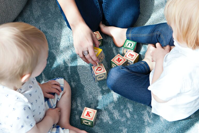 Two young children sit on a blue carpet while an adult hand helps them play with colourful wooden alphabet blocks—one of many fun things to do with your young children. The kids, dressed in light-coloured clothes, are focused on the blocks.