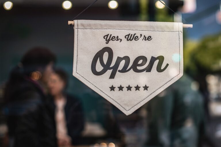 A white banner with the words "Yes, We're Open" and five stars hangs on a small business's glass door, signalling that they're open. Blurred figures and lights are visible in the background.