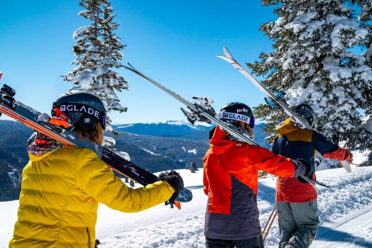 Three skiers in colourful jackets and helmets carry their skis on their shoulders, walking through snowy terrain with pine trees and mountain views—perfect for discussing tips to consider whilst choosing a good beginner ski resort.