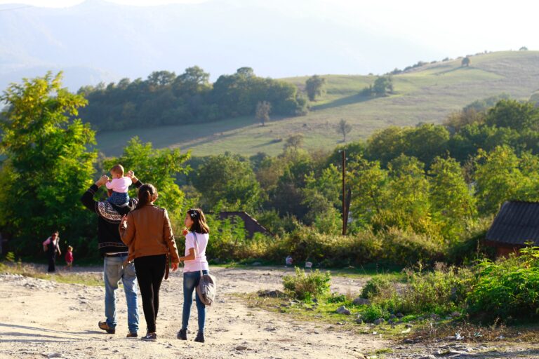 A family of four walks on a dirt path through a green, hilly countryside, juggling parenting and laundry on the go; the father carries a small child on his shoulders while the mother and another child walk beside him, surrounded by trees and sunlight.