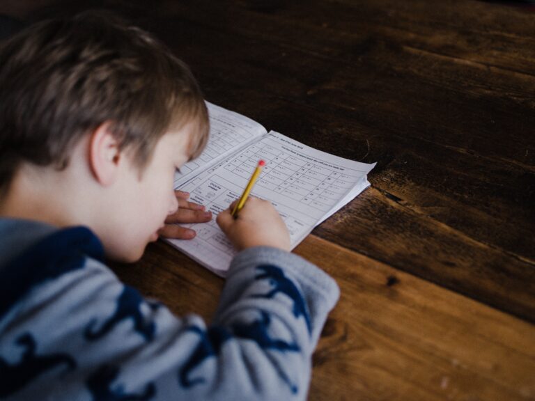 A young child wearing a dinosaur-patterned jumper writes with a pencil on a worksheet at a wooden table, focused on completing schoolwork—showcasing how to raise a hardworking child through encouragement and dedication.