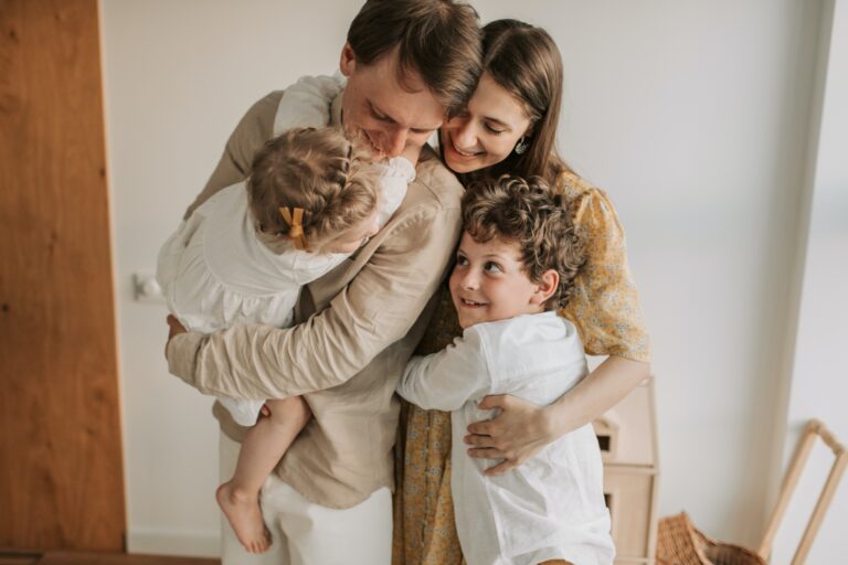 A family of four shares a group hug, smiling warmly. In their cosy home, they show how balancing your busy career with quality time with your children creates moments of togetherness in light-coloured, casual clothes.