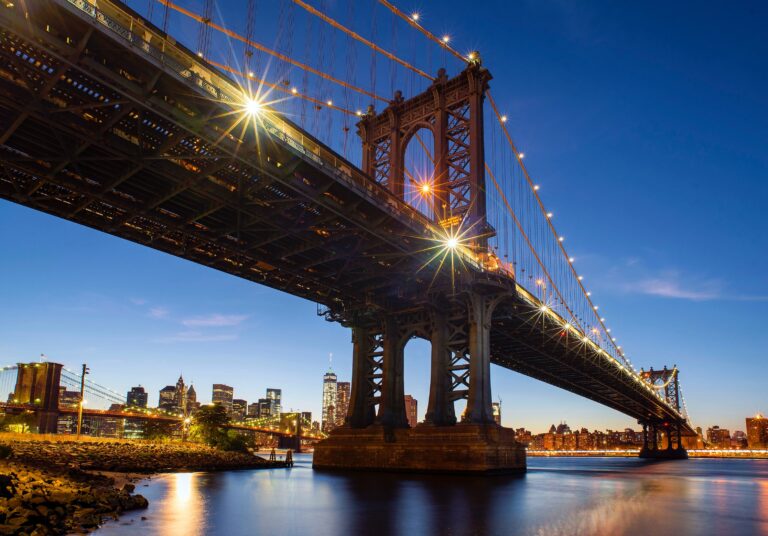 A city skyline at dusk with the Manhattan Bridge illuminated in the foreground, spanning a calm river and highlighted as one of the unique places in New York, with buildings glowing softly in the background.