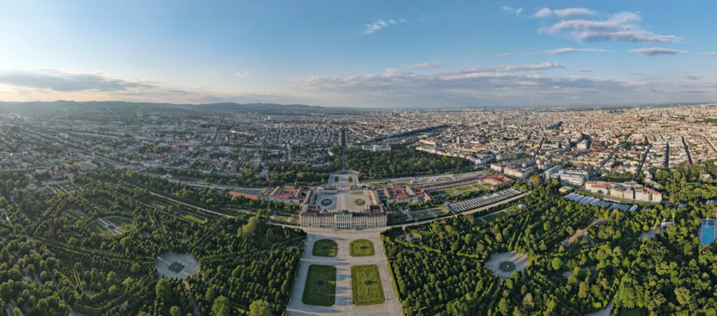 Aerial view of Schönbrunn Palace in Vienna, Austria, surrounded by symmetrical gardens and green trees, with the cityscape stretching out in the background—perfect for discovering Vienna on a budget with family-friendly holiday packages.