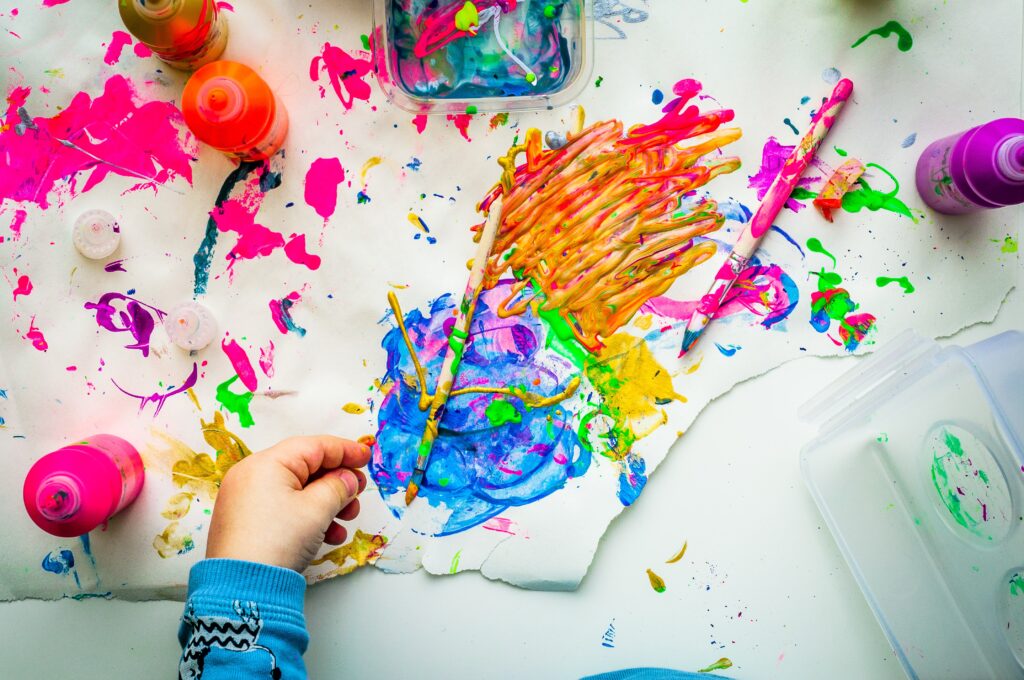 A child’s hand painting with bright colours on paper, surrounded by bottles of paint, a paintbrush, and splatters of pink, blue, green, and yellow creates a playful and creative scene perfect for family fun.